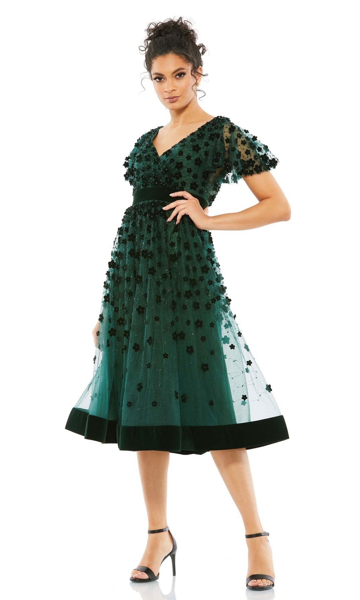 A woman in the Mac Duggal 67854 Floral Midi Wedding Guest Dress—vintage green, knee-length with short sleeves, floral appliqués, and a velvet waistband—stands in open-toe heels against a white background.